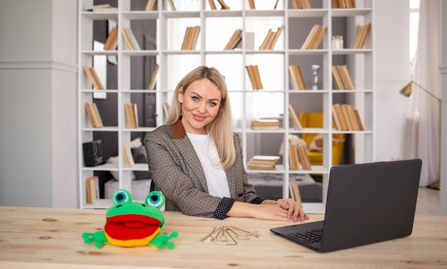Een vrouwelijke lerares zit aan een tafel in een kamer met een laptop en een gebreid kikkerspeelgoed Leraar logopedist