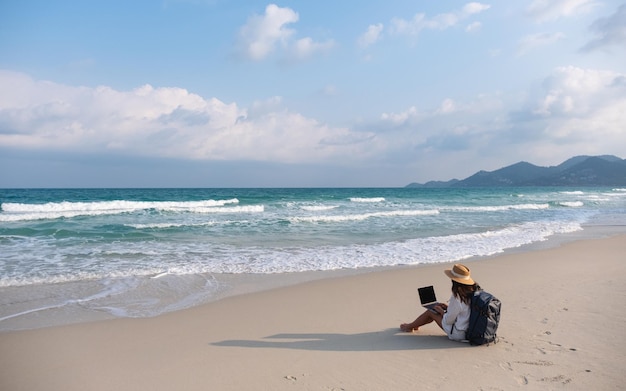 Een vrouwelijke reiziger die een laptop gebruikt en erop werkt terwijl hij op het strand zit