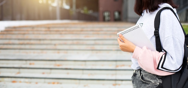 Een vrouwelijke student met een zwarte rugzak en een notitieboekje in haar handen staat op de trap voor de universiteit. Hoger onderwijs, het begin van het schooljaar, terug naar school. Copyspace