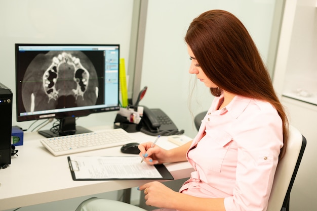 Een vrouwelijke tandarts arts zit aan een tafel, op een computer een CT-scan van de kaak. De dokter is gekleed in professionele kleding.