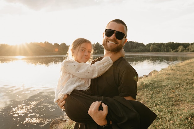 Een wazige foto van een jonge vader met een baard die zijn lachende dochter in zijn armen houdt aan de kust van het meer. Eenoudergezin op vakantie op de zonsondergang.