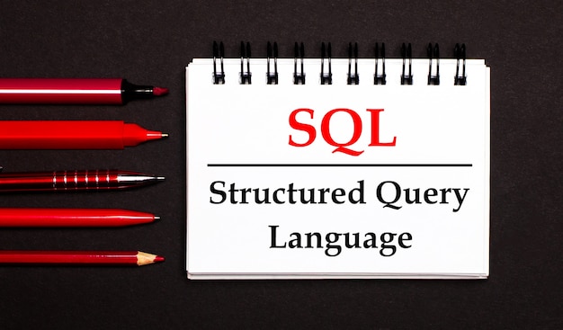 Een wit kladblok met de tekst SQL Structured Query Language, geschreven op een wit kladblok naast rode pennen, potloden en stiften op een zwarte achtergrond
