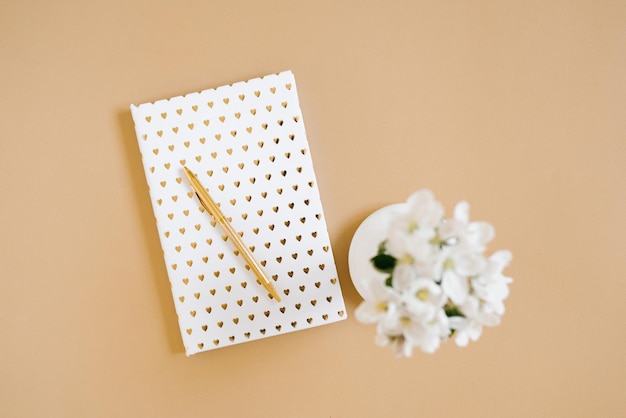 Een wit notitieboekje met een gouden pen op een beige tafel en bloemen in een defocusvaas