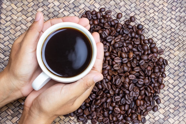 Een witte kop koffie in handen van de mens met koffiebonen naast hem.