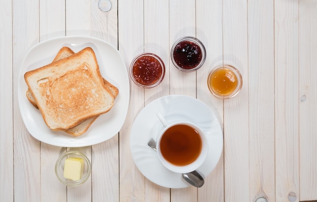 Een witte kop zwarte thee met sandwiches of toast met aardbeien, bessen en abrikozengelei of jam op witte houten tafel, bovenaanzicht, plat gelegd, ontbijtconcept met kopieerruimte