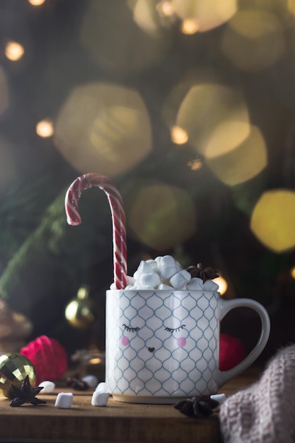 Een witte mok met een beschilderd gezicht vol marshmallowthee en een kerstsnoepstokje. Rond de kerstsfeer, lichtjes, ballen