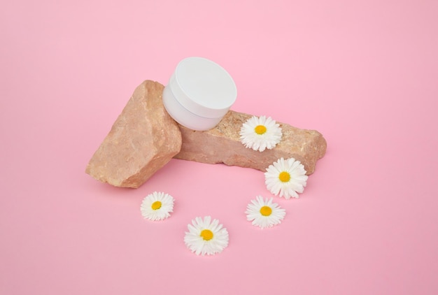 Een witte plastic pot met cosmetisch product staat op een stenen podium met witte kamillebloemen op een roze achtergrond Bio-cosmetica mock-up