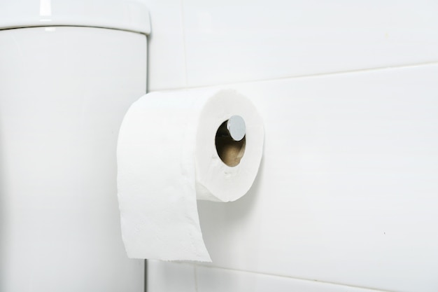 Foto een witte rol van zacht toiletpapier netjes opknoping op chromen houder op een witte badkamer muur. detailopname