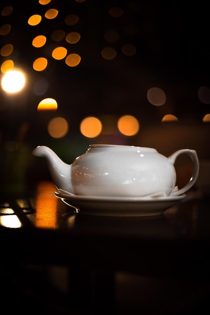 Een witte ronde theepot staat op een houten bar in een uitgaanscafé. Tegen de achtergrond van gouden cirkels van gele kerstbokehslingers. Zachte focus. Nachtthee.