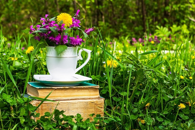 Een witte theekop met een boeket wilde bloemen op een stapel oude boeken in een weiland met groen gras op een zonnige lente- of zomerochtend