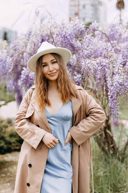 Een zacht meisje in een jurk en een modieuze hoed loopt en geniet van de geur van bloemen in het park.