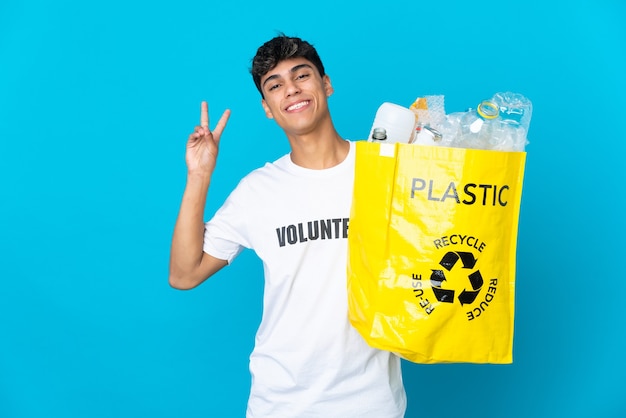 Een zak vol plastic flessen vasthouden om te recyclen op een blauwe achtergrond die glimlacht en een overwinningsteken toont