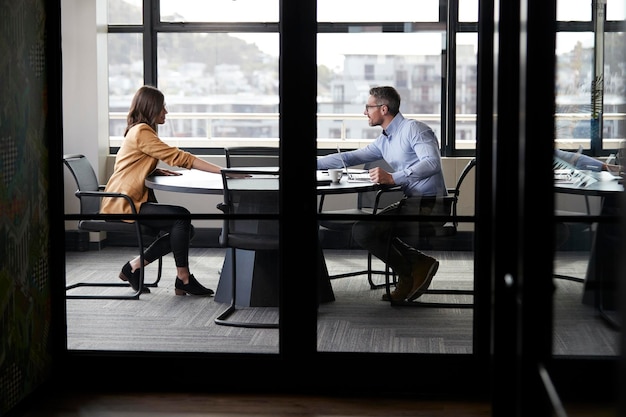 Een zakenman en een jonge vrouw ontmoeten elkaar voor een sollicitatiegesprek van volledige lengte gezien door een glazen wand