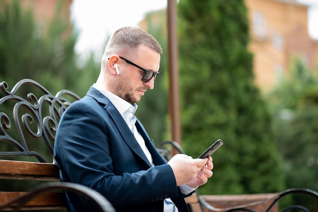 Een zakenman in een donkere zonnebril met een telefoon in zijn handen zit op een bankje en praat aan de telefoon