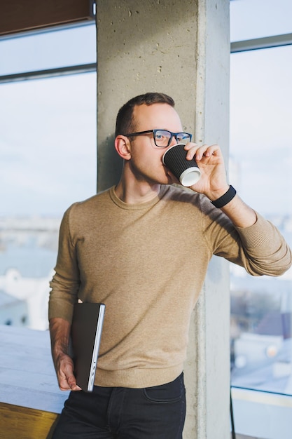 Een zakenman in glazen en stijlvolle kleding drinkt koffie in een moderne werkruimte Een mannelijke freelancer met een laptop werkt op afstand