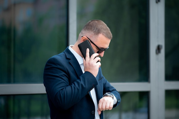 Een zakenman staat buiten het bedrijfsgebouw, praat aan de telefoon en kijkt op het horloge