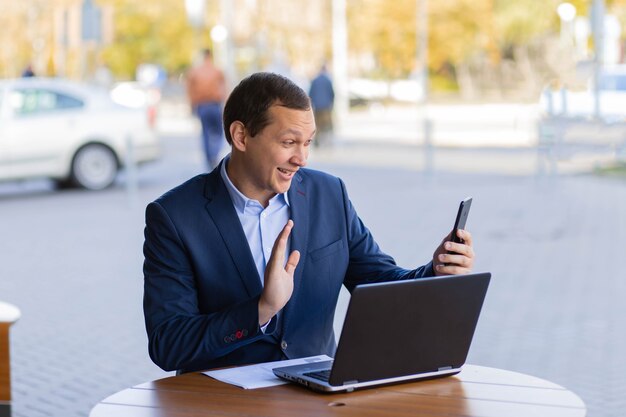 Foto een zakenman zit aan een tafel in een café op straat en zwaait met zijn hand tijdens een videogesprek naar zijn mobiele telefoon tijdens een pauze van zijn werk