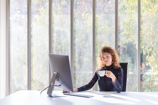 Een zakenvrouw heeft een lachend gezicht en drinkt 's ochtends koffie op haar kantoor met een computer.