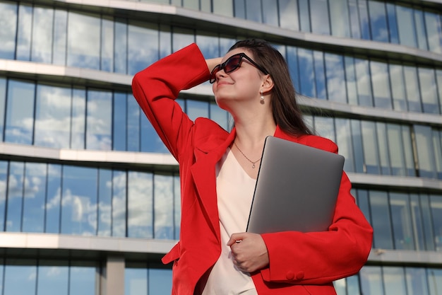 Een zakenvrouw in een jas poseert met een laptop tegen de achtergrond van een kantoorgebouw