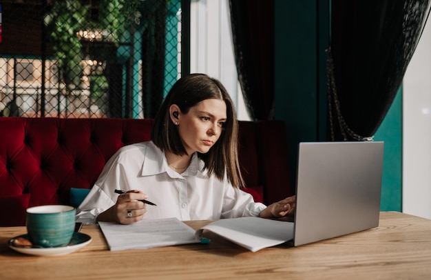 Een zakenvrouw met een draadloze koptelefoon zit aan een tafel in een café met documenten en werkt op een laptop