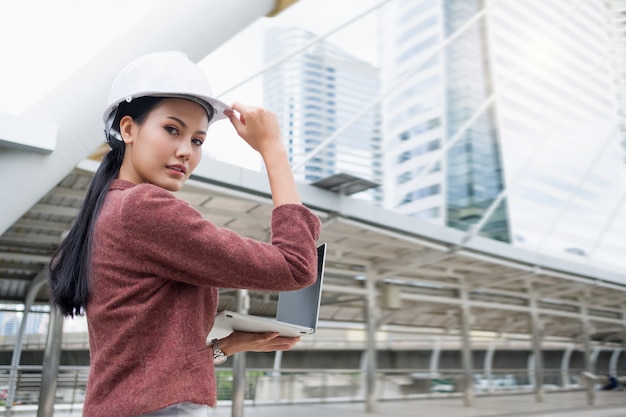 Een zelfverzekerde Aziatische werkende vrouw draagt een helm en werkt op een laptop terwijl je buitenshuis staat.