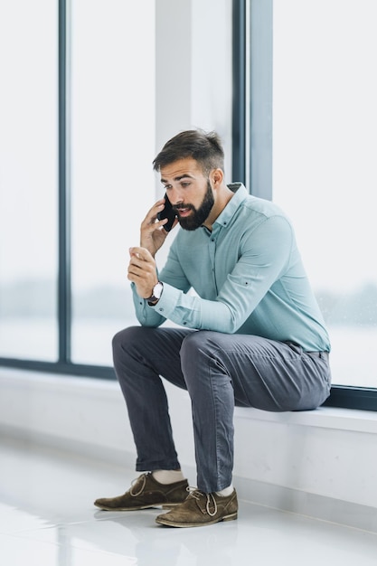 Een zelfverzekerde zakenman die tijdens de pauze op een smartphone praat terwijl hij bij het raam in de kantoorhal zit.