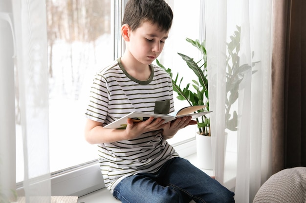 Een zesjarige jongen leest een boek terwijl hij bij het raam zit Thuisonderwijs