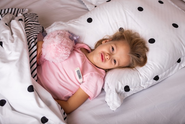 Een ziek kind ligt in bed met een zacht stuk speelgoed en meet de temperatuur met een thermometer