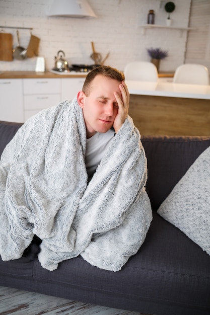 Een zieke man zit thuis op een grijze bank met een witte deken. Ziekte, bescherming, coronavirus, ziekte, griep.