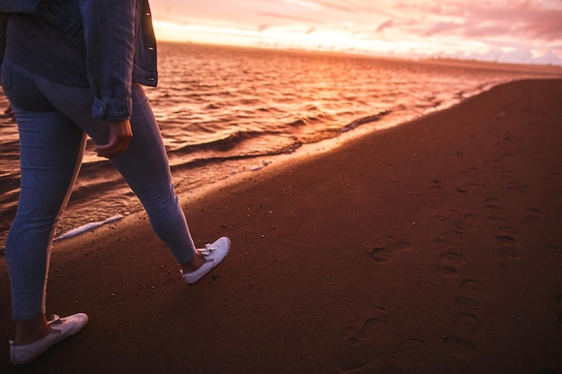 Een zijaanzicht een meisje loopt alleen op het strand bij de kleurrijke zonsondergang na de regen Benen in de witte schoenen Oranje karmozijnrode paarse kleuren