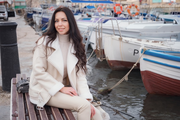 Een zorgeloze blanke vrouw in beige kleding genietend van het uitzicht op de zee op een warme, winderige dag. portret voor een zeebaai met boten op de pier