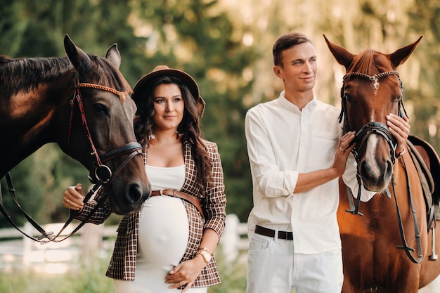 Een zwanger meisje in een hoed en haar man in witte kleren staan naast paarden in het bos in de natuur. Stijlvolle zwangere vrouw met een man met paarden. Familie.