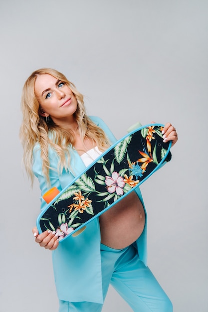 Een zwanger meisje in turquoise kleding met een skateboard in haar handen op een grijze achtergrond