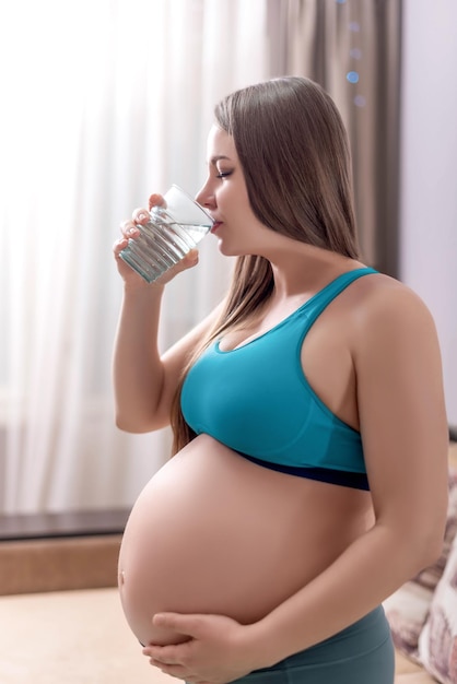 Een zwanger meisje met een glas in haar hand drinkt water in sportkleding