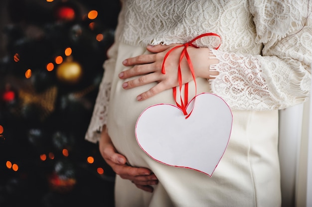 Een zwangere vrouw houdt haar armen om de ronde buik. bevat een versierd hart met een lint. uitzicht op de maag zwangere vrouw. wachten op een baby. gelukkig familieportret, concept van een vakantie met het gezin.