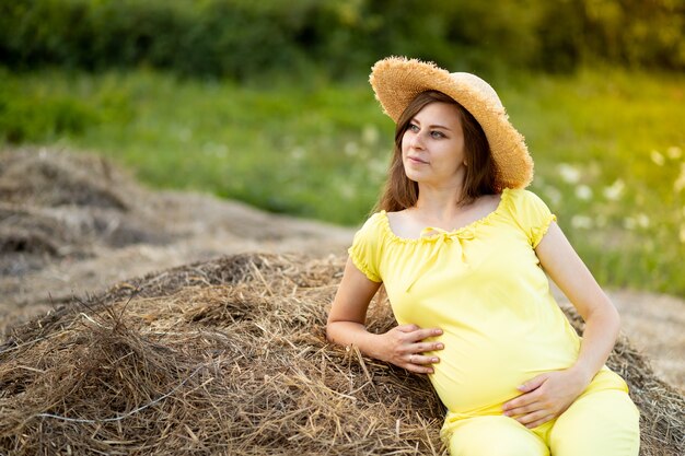 Een zwangere vrouw in donkere kleren en een hoed zit in een veld op stro in de zomer