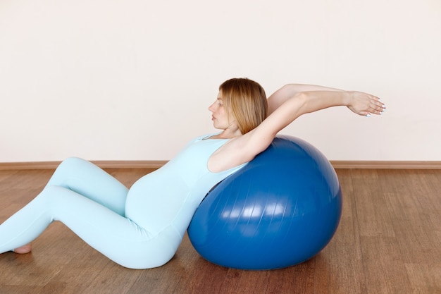 Een zwangere vrouw in een blauwe jumpsuit strekt zich uit op een fitnessbal in een yogastudio. yoga voor zwangere vrouwen