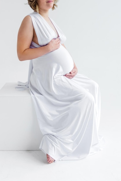 Een zwangere vrouw in een lange witte zijden jurk knuffelt haar buik en poseert op een witte achtergrond zittend op een witte kubus