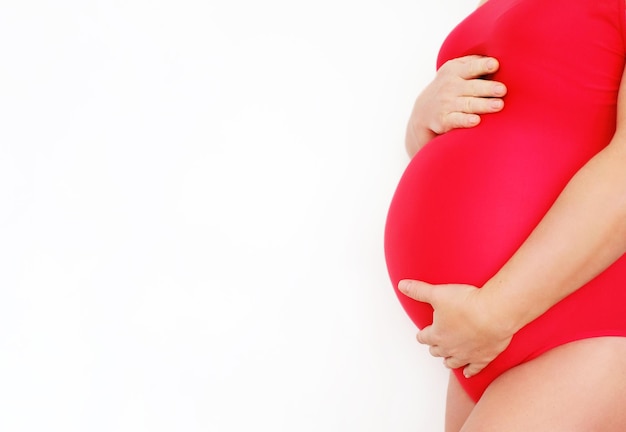 Een zwangere vrouw in een rood badpak strijkt over haar buik en poseert op een witte achtergrond Close-up van de buik