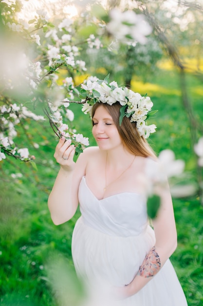 Een zwangere vrouw in een witte jurk en een bloemenkrans. Zwangere vrouw in een bloeiende botanische tuin dichtbij bloeiende sakura en appelbomen
