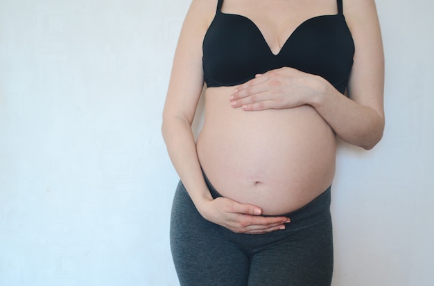 Een zwangere vrouw staat tegen een witte muur en streelt haar buikconcept van lichaamshouding en zwangerschap