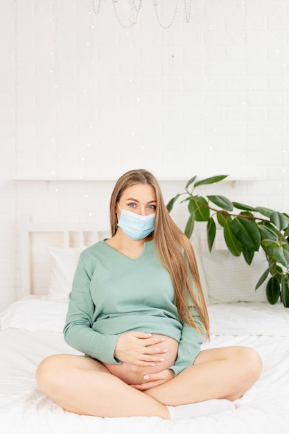 Een zwangere vrouw zit in een medisch beschermend masker op het bed en raakt haar grote buik thuis aan, het concept van zwangerschap en wachten op de geboorte van een baby en een lange termijn