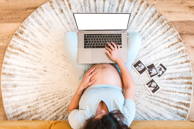 Een zwangere vrouw zit op het tapijt en gebruikt een laptop om te winkelen of om echografiebeelden te maken