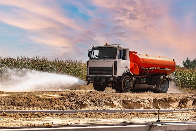 Een zware vrachtwagen giet water op de weg op een bouwplaats waar bestratingsplaten worden gelegd Stofverwijdering milieubescherming Irrigatie van de weg tegen stof en bodemverdichting in de bouw