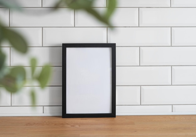 Een zwart frame met een leeg doek tegen een witte tegelmuur en op een houten tafel met vaag groen gebladerte