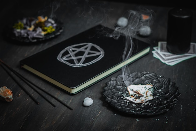 Een zwart metalen schotel met verbrand papier, een leren boek met een magisch teken rituele stenen en een kaars op een zwarte achtergrond