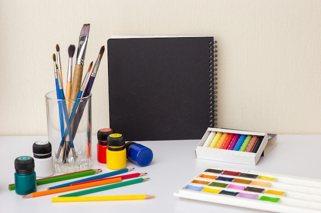 Een zwart schetsboek op een witte tafel met tekenbenodigdheden en borstels