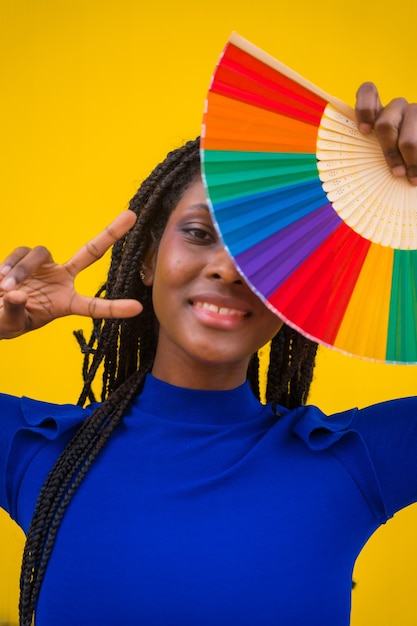 Foto een zwarte etnische vrouw met een regenboog lgbt-fan op een gele achtergrond die het overwinningssymbool maakt