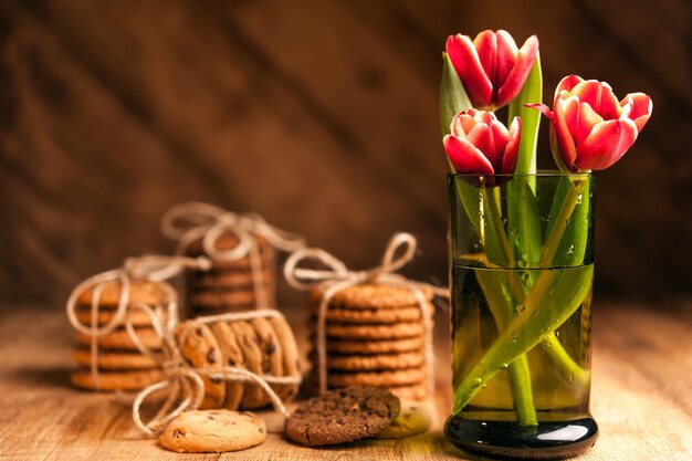 Eenvoudig rustiek stilleven met bloeiende rode tulpen in een groen glas en stapelt verschillende koekjes op een donkere houten tafel. Fine art rustieke achtergrond. Detailopname.