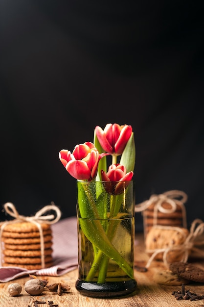 Eenvoudig rustiek stilleven met bloeiende rode tulpen in een groen glas en stapelt verschillende koekjes op een donkere houten tafel. Fine art rustieke achtergrond. Detailopname.